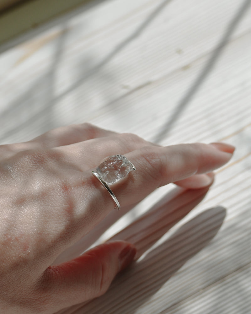 A rhodium‑plated ring with transparent glass forms reminiscent of melting glaciers, gently held between upper and lower bands that wrap around the finger.
