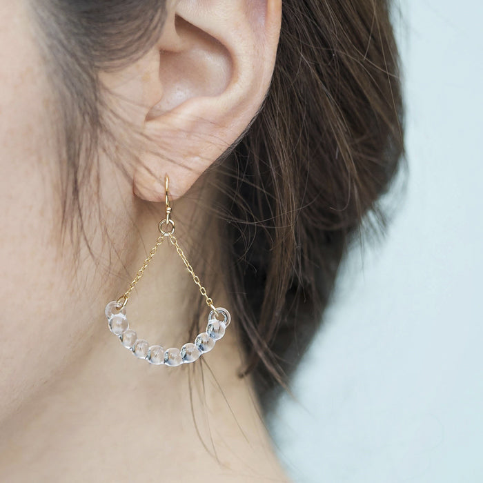 Handmade glass arc pieces resembling gathered water droplets, suspended at both ends by twin gold‑plated chains from a hook.